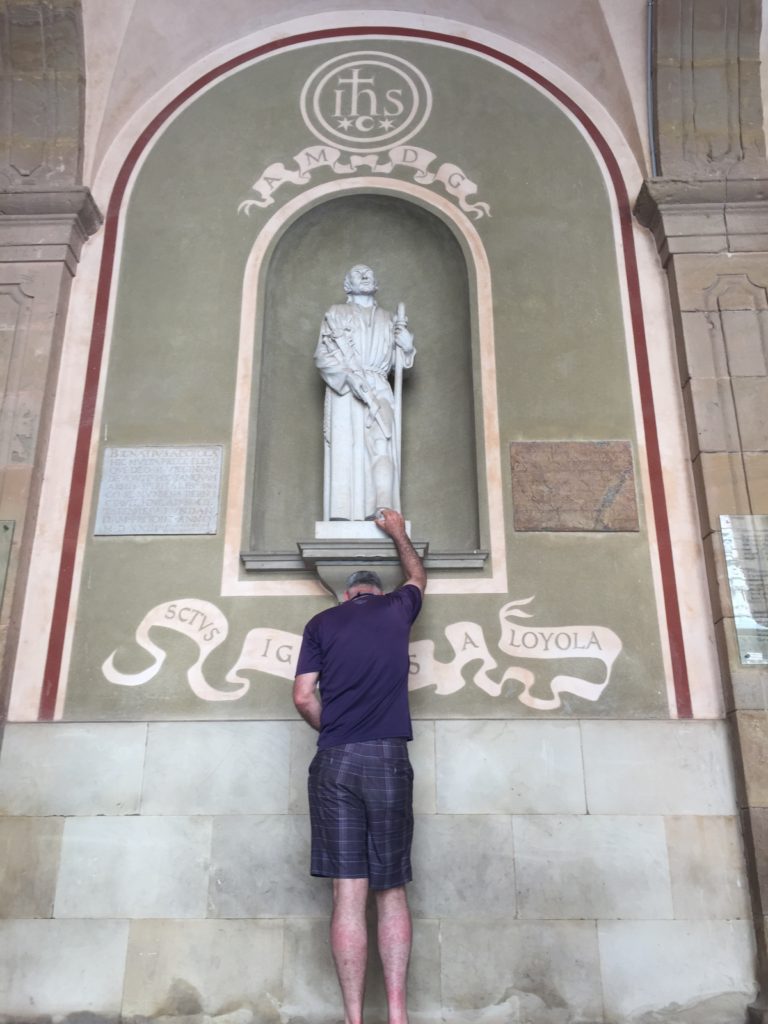 Man standing under statue of St. Ignatus reaching hand up to touch his toe.