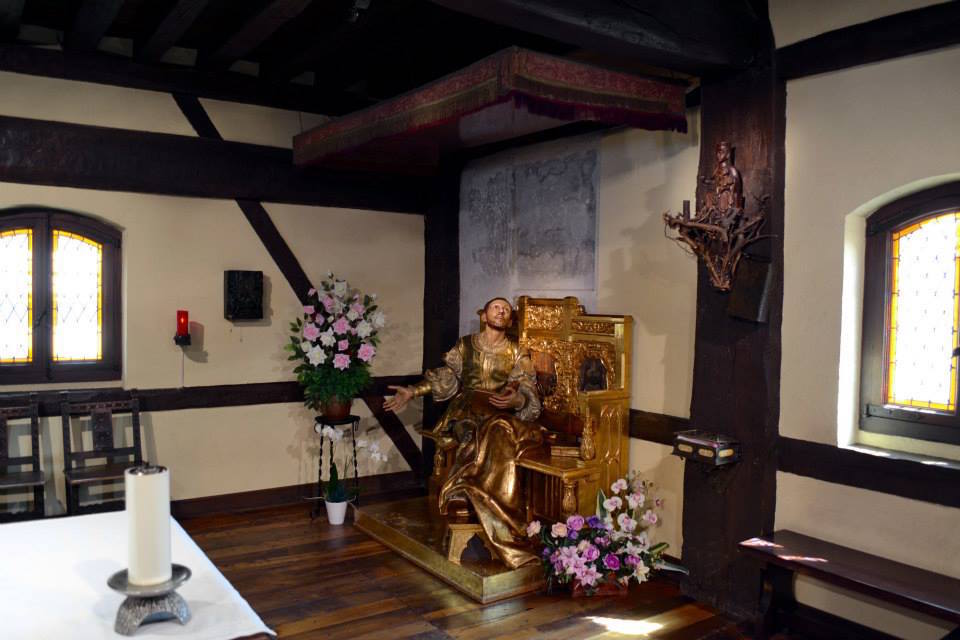 Bronze statue of Ignatius of Loyola seated in a chair in midst of simple room with wood floor.