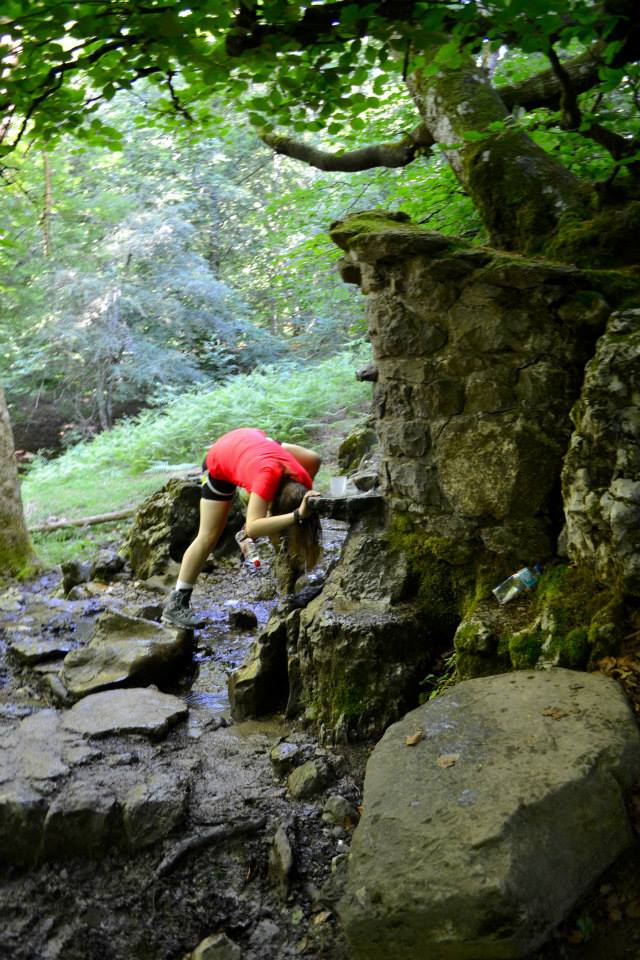 Woman in red shirt bending to place head under spring pouring over rock